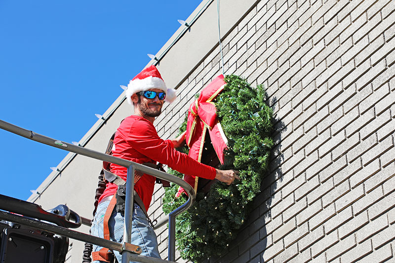 man in a Santa hat hanging a large wreath from a tree bucket
